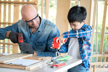 A senior carpenter and boy wearing goggle in the modern wood workplace. A man looking at boy who used screwdriver. Young child concentrate and learning from teacher. Education and learning concept