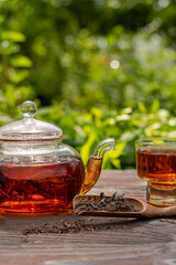 Steamed glass teapot, elegant cup. Bamboo spoon with tea, wooden table. Cropped vertical view, outdoor, picnic, brunch. Floral background in blur.