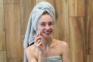 Young woman makes a facial massage with a gouache scraper in home bathroom. Beautiful girl wearing bath towel on head portrait. Skin care after cleansing