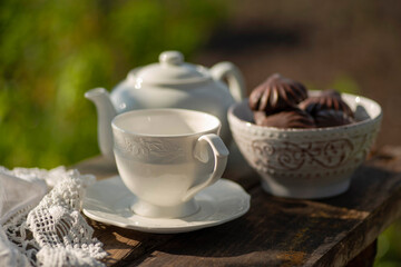 Elegant cup, lace tablecloth, teapot, marshmallow in chocolate, wooden table. Outdoor breakfast, picnic, brunch, spring mood. Soft focus