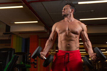 Fototapeta na wymiar A male athlete with a bare torso and red shorts stands in the gym holding dumbbells and looking away against the backdrop of sports equipment. High quality photo