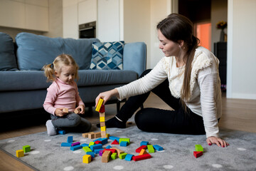 Naklejka premium Mother and daughter playing wooden cubes on the floor of the living room at home. Activities with children inside, early childhood development