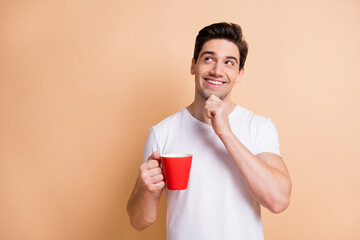 Portrait of cheerful handsome guy hand on chin look empty space hold coffee isolated on beige color...