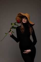Pregnant girl in black tight dress. In a big hat. With a rose in my hand. Supporting the abdomen. Grey background.