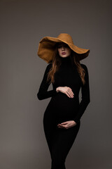 Pregnant girl in black tight dress. In a big hat. Hands on belly. Looking down. Grey background.
