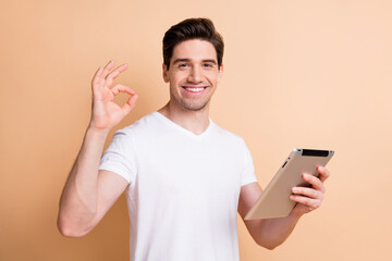 Portrait of half turned cheerful guy arm hold tablet fingers show okey symbol isolated on beige color background