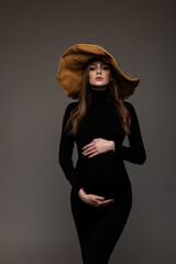 Pregnant girl in black tight dress. In a big hat. Hands on belly. Grey background.