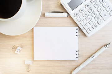 A notebook with a white blank sheet on a wooden table next to a white coffee cup and a white calculator with a pen. Business concept