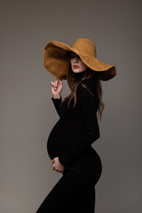 Pregnant girl in black tight dress. In a big hat. Profile. Hand holding hat. Grey background.
