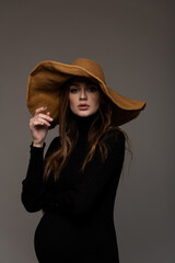 Portrait pregnant girl in black tight dress. In a big brown hat. Hand to face. Grey background.