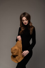 Pregnant girl in black tight dress. Big hat in hand. Looking down. Grey background.