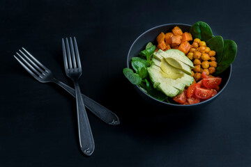 Avocado poke bowl with cherry tomatoes, chickpeas, sweet potato, spinach and lamb's lettuce on black dark background.Closeup view of vegetarian Buddha bowl ,healthy food concept.