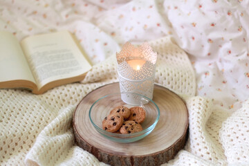 Bowl of chocolate chip cookies, candle holder with lit candle, wooden tray, open book and knitted blanket at home. Selective focus. 