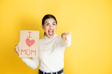 Beautiful woman celebrating mothers day holding poster love mom message pointing with finger surprised ahead, open mouth amazed expression, something on the front