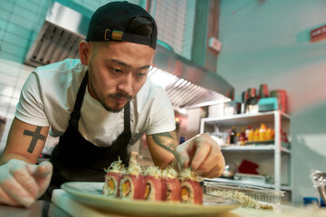 Portrait of experienced sushi chef carefully adding final touch with dedication to his delicious sushi set - obrazy, fototapety, plakaty
