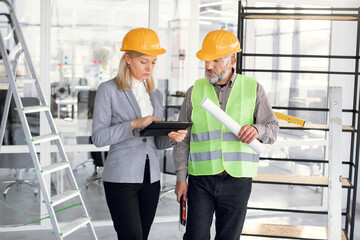 Qualified female designer using digital gadget while standing near bearded contractor indoors. Man and woman wearing protective helmets at work. Control of working process.