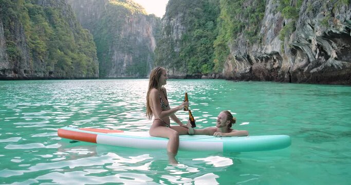 Happy young romantic caucasian couple male and female sitting on stand up paddle board enjoying summer vacation having fun celebrating summertime holiday with smiling face drinking beer and kissing.
