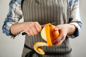 Woman peeling fresh orange with knife on light grey background, closeup