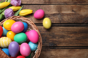 Fototapeta na wymiar Colorful Easter eggs in wicker basket and tulips on wooden table, flat lay. Space for text