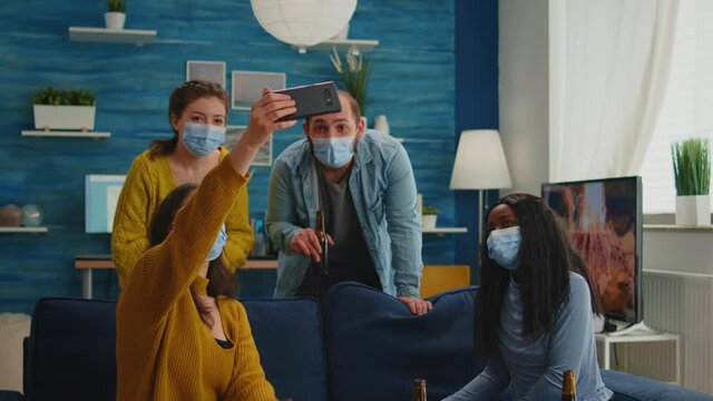 Cheerful happy mixed race friends taking selfie wearing face mask keeping social distancing against spread of virus sitting on couch in living room. Diverse people socializing keeping social distance