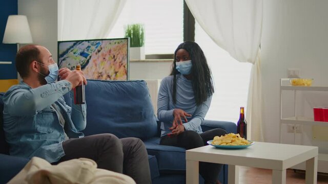 Mixed friends with protection mask, talking during new normal party keeping distance sitting on sofa drinking beer and eating snaks. Group of multi ethnic people socializing and enjoying free time