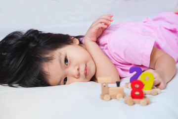 Obraz na płótnie Canvas Adorable Asian girl lying down playing wooden figures toys on mattress. Child looks at the camera. Little kid relaxing day. Children are happy and feel safe in the home. Cute baby 3-4 years old.