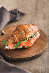 One croissant sandwich with salmon, ricotta cheese and arugula on a wooden board with a napkin on a dark background.