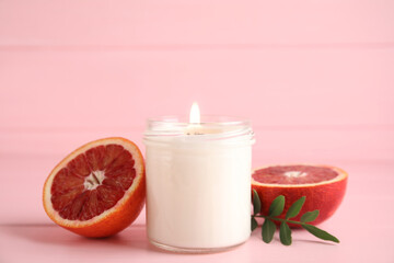 Scented candle with burning wooden wick and cut grapefruit on pink table