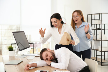 Young women popping paper bag behind their sleeping colleague in office. Funny joke