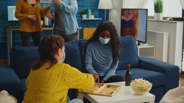 African woman competing with friends playing backgammon wearing face mask keeping social distancing during social pandemic with covid19 drinking beer in living room. Enjoying board games in outbreak