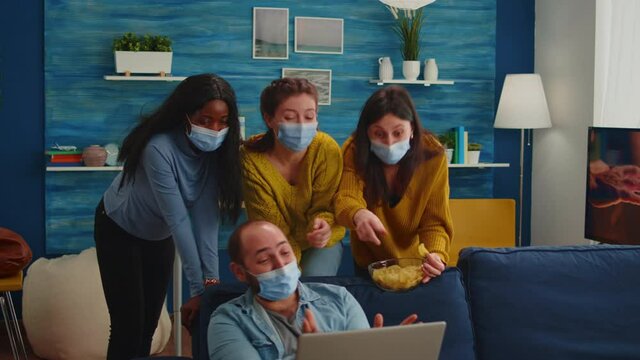 Multi ethnic friends wearing face mask eating snacks using laptop keeping social distancing to prevent coronavirus spread during global pandemic having fun in home living room. Conceptual image.
