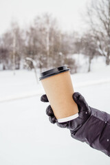 A man's gloved hand holds a cup of coffee. A mockup cup of coffee. Hot coffee to take away. Cappuccino with almond milk on the street. Craft cup with lid in hand