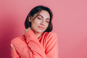Smiling young woman 20s in casual clothes isolated on pink background studio portrait. Love concept. Mock up copy space. Keeping eyes closed holding hands crossed hugging herself