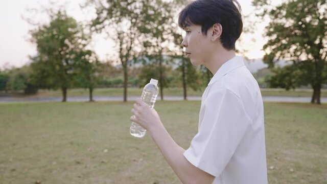 A Young Thirsty Man Drinking A Pure Water While Walking Inside Green Park On Weekend,  Holding A Reusable Clean Mineral Bottle Water, Lifestyle Healthcare Concept, Ecosystem Caring, Beauty Sunset Time