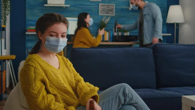 Lady looking at camera enoying spending time with friends in living room keeping social distancing wearing face mask to prevent coronavirus spreading. during global outbreak holding beer bottle.