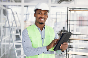 Smiling afro-american engineer in construction hardhat working on digital tablet. Positive man in uniform using modern gadget at work. Business person.