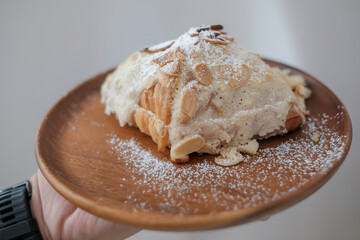  Fresh croissant breakfast. Croissant with almond, classic traditional French breakfast on white plate, top of icing sugar and almond slide. Light brown wood background.