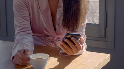 Young caucasian female wearing velvety rose robe with a cup of hot tea or coffee in the hand. The woman is navigating in her phone. Sunny day. Closeup no face high-resolution image.