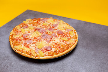 Simple Hawai pineapple ham oven pizza with crunchy crest and golden orange tint on modern rustic gray surface. Studio food still life against a seamless yellow background