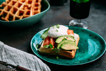 Belgian waffles with salmon, poached egg, avocado and cream cheese on a turquoise plate. Healthy breakfast at the table