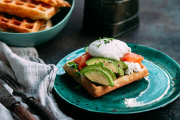 Belgian waffles with salmon, poached egg, avocado and cream cheese on a turquoise plate. Healthy breakfast at the table