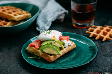 Belgian waffles with salmon, poached egg, avocado and cream cheese on a turquoise plate. Healthy breakfast at the table