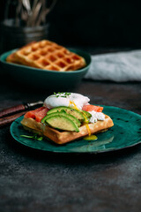 Belgian waffles with salmon, poached egg, avocado and cream cheese on a turquoise plate. Healthy breakfast at the table