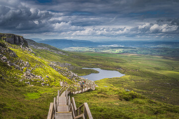 People hiking on steep stairs of wooden boardwalk in Cuilcagh Mountain Park. View on lake and valley below with dramatic sky, Northern Ireland - obrazy, fototapety, plakaty