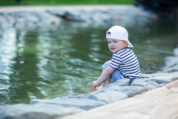 Baby in a white cap sitting by the pond touches cold water with his leg and admires