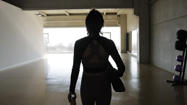 Tracking shot, rear view of caucasian slim Woman, with a fitness mat and water bottle, entering in a yoga center to start pilates fitness routine. Early morning, Big windows, slow motion.