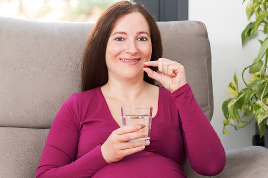Happy Pregnant Woman Taking A Red Pill At Home