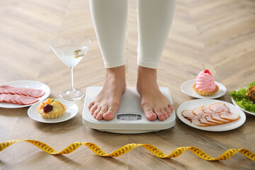 Food, alcohol left after holidays and woman with measuring tape standing on scales indoors,...