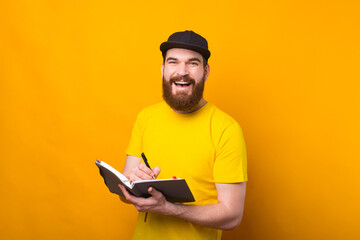 Cheerful young bearded hipster man writing in agenda and smiling