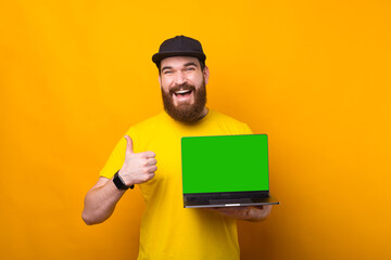 Joyful young bearded hipster man showing thumb up and green screen on laptop - Powered by Adobe